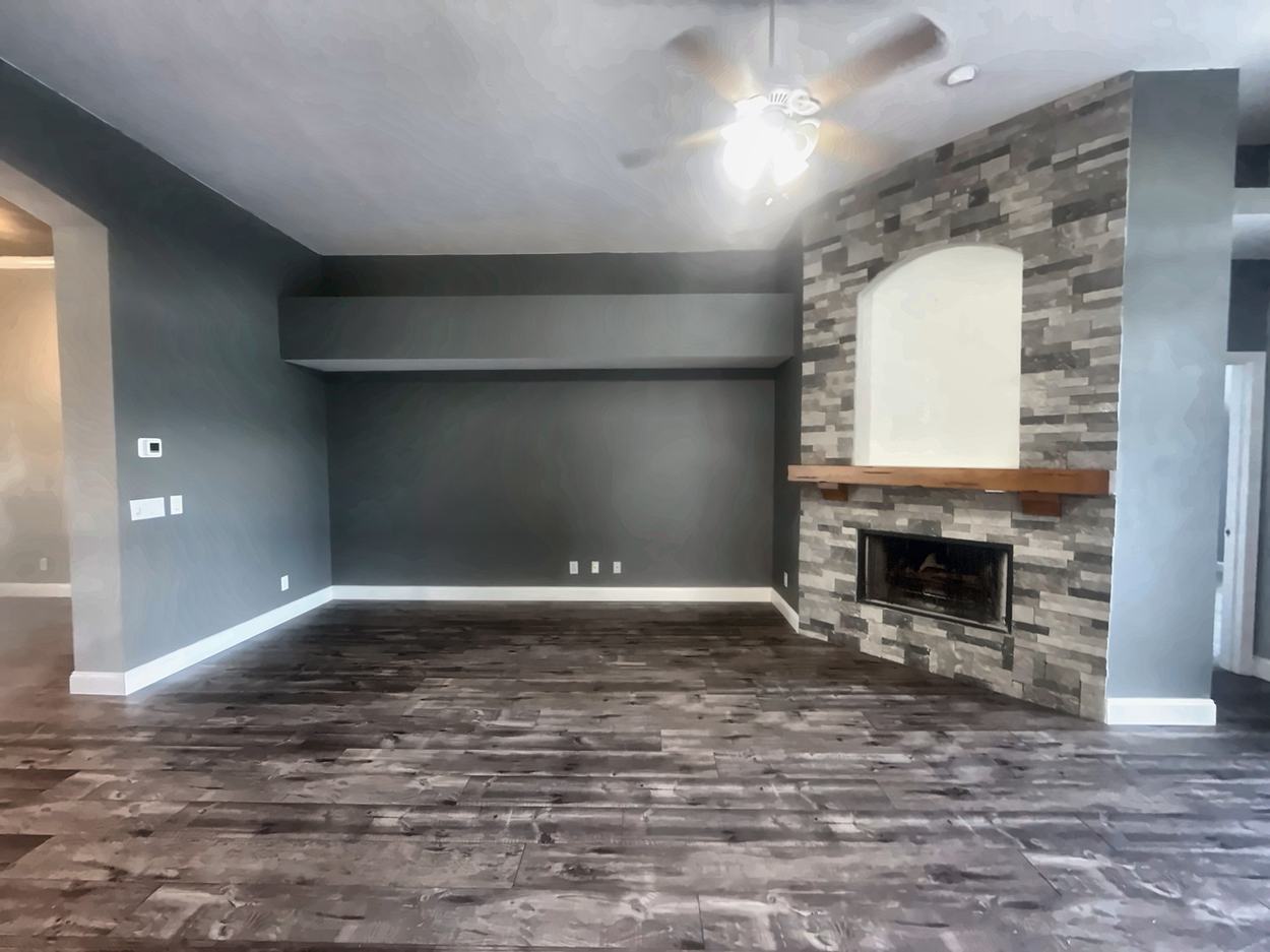 Great room before renovation: a stacked stone fireplace with an arched plaster niche and rustic timber mantel, under a dropped soffit, with grey walls and a ceiling fan
