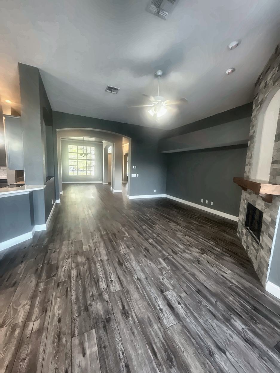Open plan before renovation, looking from the kitchen counter down the length of the living room toward the dining room, with grey walls and the stone fireplace at right