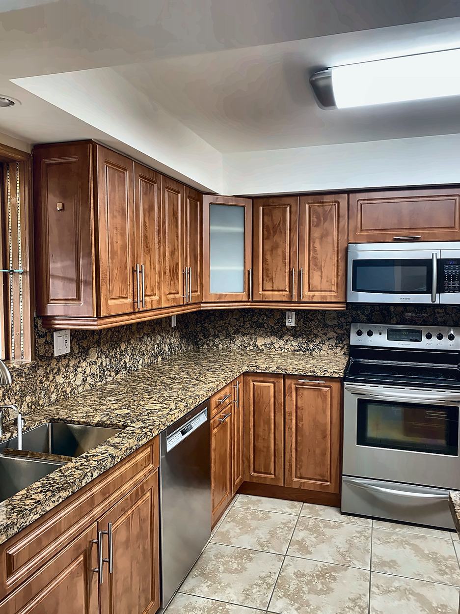The kitchen before renovation: raised-panel cherry cabinets, speckled granite counters and backsplash, stainless appliances and beige ceramic floor under a boxed fluorescent light