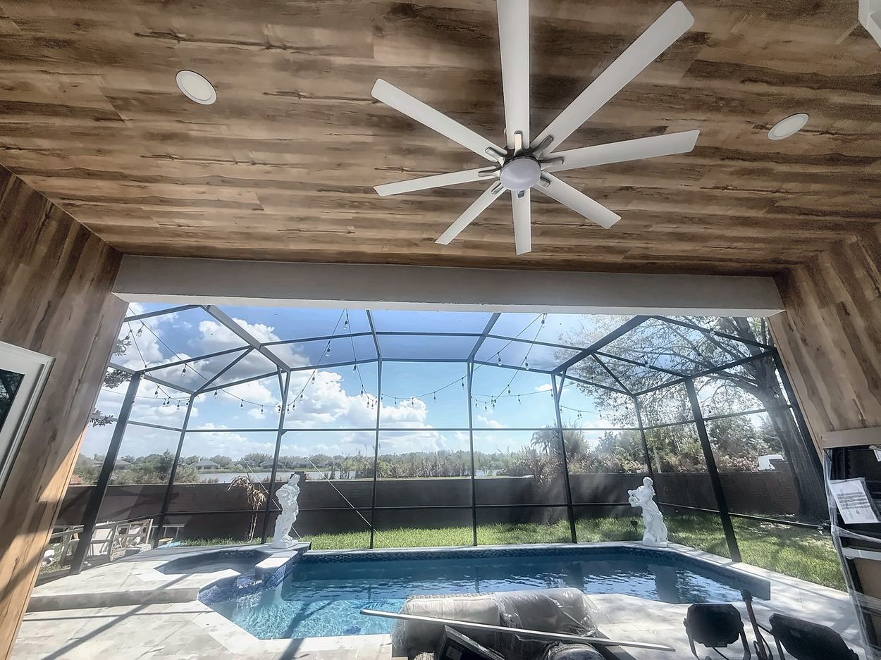 Covered lanai with a wood-plank ceiling and large fan, opening through a screen enclosure onto the pool and the lake beyond